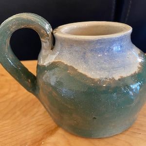 May include: A small, green and white ceramic pitcher with a handle. The pitcher has a glazed finish and a rustic look.