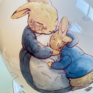 May include: A ceramic pitcher with a cream-colored background. The pitcher features a detailed illustration of two rabbits embracing. One rabbit wears a blue jacket, and the other is in a yellow-brown color. Floral patterns are visible on the side.