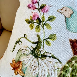 Pu&ograve; includere: Cuscino decorativo color crema con ricamo a tema autunnale. Presenta una zucca azzurra, fiori rosa, foglie verdi e un uccello blu. Il cuscino ha una superficie strutturata e un'estetica accogliente e stagionale.