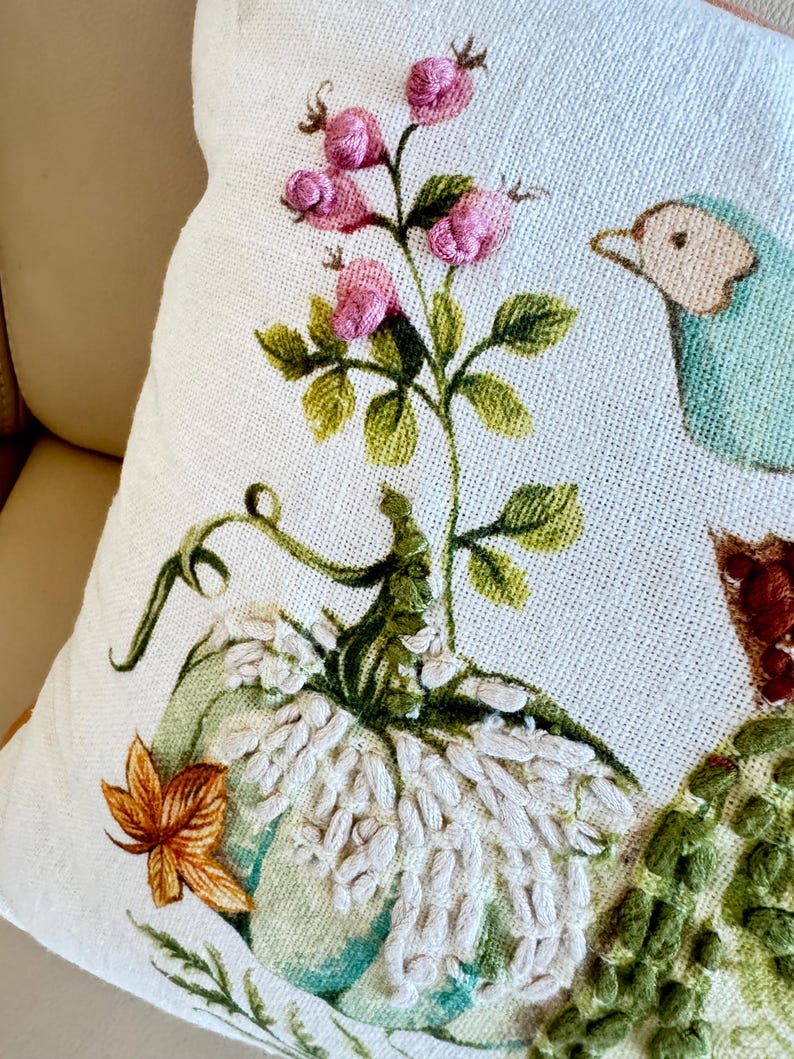 Pu&ograve; includere: Cuscino decorativo color crema con ricamo a tema autunnale. Presenta una zucca azzurra, fiori rosa, foglie verdi e un uccello blu. Il cuscino ha una superficie strutturata e un'estetica accogliente e stagionale.