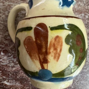 May include: A small, hand-painted ceramic pitcher with a cream-colored base. The pitcher features brown, green, and blue floral designs. It has a handle and a spout, and is approximately 4 inches tall.
