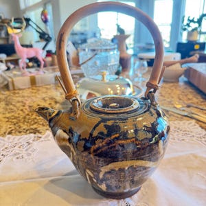 May include: A ceramic teapot with a wooden handle. The teapot has a brown, blue, and white mottled glaze. The handle is made of a curved piece of wood. The teapot is sitting on a white lace tablecloth.