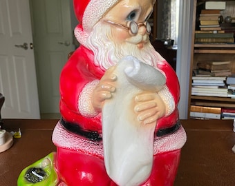 May include: A vintage Santa Claus figurine made of plastic. He is wearing a red suit with white trim and a white beard. He is holding a white sack in his hands. He is sitting on a green bag.