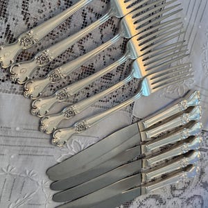 May include: A collection of silver forks and knives arranged on a white lace tablecloth. The silverware features ornate floral detailing on the handles. The forks are arranged in a fan shape, and the knives are parallel.