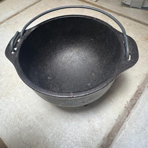 May include: A black cast iron Dutch oven with a curved handle. The pot has a rounded shape with three small feet. The handle is made of a dark gray metal. The interior of the pot is smooth and dark.