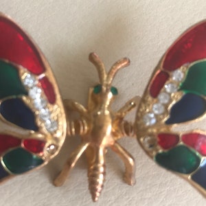Puede incluir: Un broche de mariposa de tono dorado con esmalte rojo, verde y azul y brillantes transparentes.