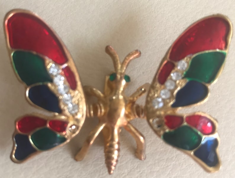Puede incluir: Un broche de mariposa de tono dorado con esmalte rojo, verde y azul y brillantes transparentes.