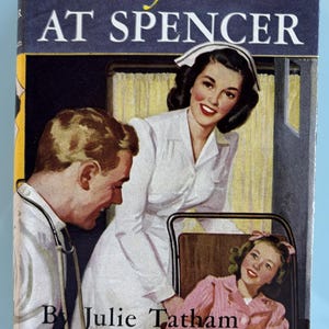May include: A book cover featuring a nurse in a white uniform, a doctor with a stethoscope, and a young girl in a pink shirt and yellow blanket. The book title is "Cherry Ames at Spencer" by Julie Tatham.