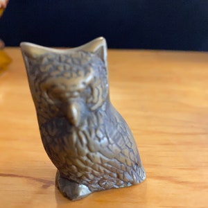 May include: A small, detailed brass owl figurine. The owl is sitting with its wings folded and has a textured surface.