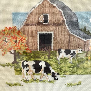 May include: Cross-stitch artwork depicting a rural farm scene. A brown barn with a gray roof and a black door is in the background. Two cows with black and white markings graze in a green field. An autumn tree with red and orange leaves is on the left.