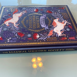 Op de afbeelding: Hardcover boek getiteld "The Magical Unicorn Society Official Handbook". De donkerblauwe kaft heeft gouden details en toont illustraties van eenhoorns, bloemen en bladeren. De titel staat ook op de rug van het boek.