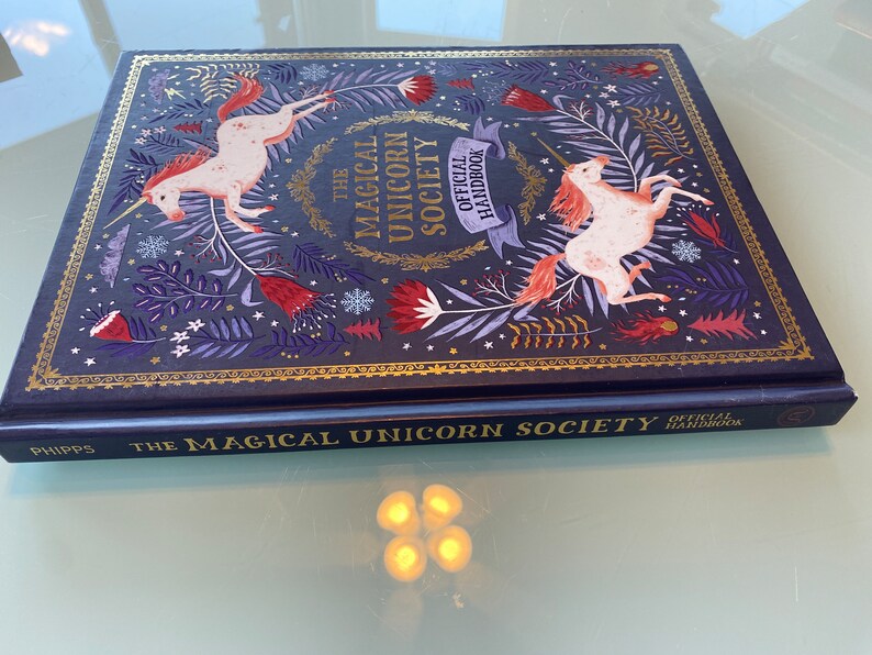 Op de afbeelding: Hardcover boek getiteld "The Magical Unicorn Society Official Handbook". De donkerblauwe kaft heeft gouden details en toont illustraties van eenhoorns, bloemen en bladeren. De titel staat ook op de rug van het boek.