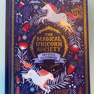 Op de afbeelding: Een hardcover boek getiteld "The Magical Unicorn Society Official Handbook". De omslag is donkerblauw met gouden accenten en illustraties van eenhoorns, bloemen en bladeren. De eenhoorns zijn wit met roze manen en staarten.