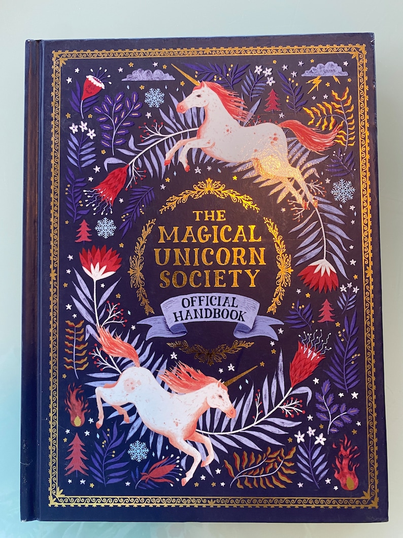Op de afbeelding: Een hardcover boek getiteld "The Magical Unicorn Society Official Handbook". De omslag is donkerblauw met gouden accenten en illustraties van eenhoorns, bloemen en bladeren. De eenhoorns zijn wit met roze manen en staarten.