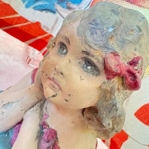 May include: A vintage figurine of a young girl with blue eyes and a red bow in her hair. The figurine has a distressed, aged appearance with peeling paint, suggesting it is an antique or a piece with a weathered aesthetic. The girl's face is detailed.