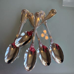 May include: Six silver spoons with a shell design on the handle. The spoons have a reflective surface, and the handles are connected with a red material. The spoons are arranged in a fan shape.