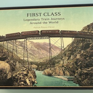 May include: A book titled "First Class: Legendary Train Journeys Around the World" by Patrick Poivre d'Arvor. The cover features a vintage photograph of a train crossing a bridge over a river in a mountainous landscape.