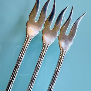 May include: Three silver-toned metal cocktail forks featuring two long, curved tines and a decorative handle with a raised beaded texture along the edges. The forks have a metallic sheen and a distinct, slender design with a scalloped base.