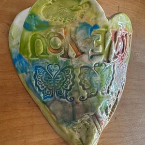 May include: A green, blue, and orange heart-shaped ceramic wall hanging with the word "love" written on it. The heart is decorated with butterfly designs.