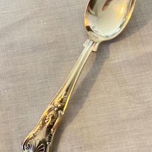May include: A silver spoon with an ornate handle featuring floral and scroll designs. The spoon has a polished, reflective surface and is set against a light-colored background. The spoon is likely for serving or eating.
