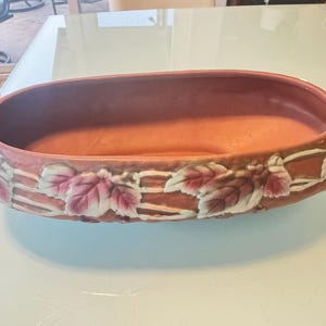 May include: A terracotta-colored, oval-shaped planter with a decorative border of leaves and vines. The planter has two small handles and measures approximately 14 inches long.