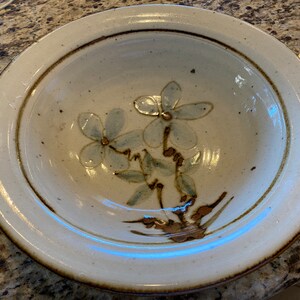 May include: A white ceramic bowl with a brown rim and a floral design featuring two blue flowers with brown centers. The bowl has a speckled glaze.