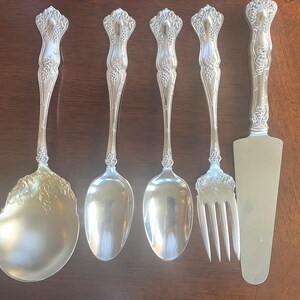 May include: A set of five silver-toned serving utensils featuring ornate floral and grape vine embossed handles. The collection includes a large serving spoon, two smaller spoons, a four-tined serving fork, and a flat triangular cake server.
