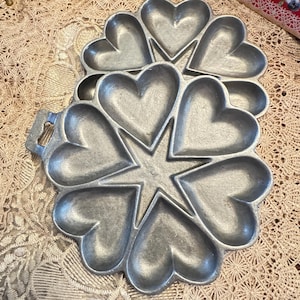May include: Two cast iron heart-shaped cake pans. Each pan has six heart-shaped molds surrounding a central star. The pans are made of a silver-colored metal and have a handle on one side.