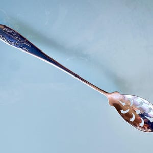 May include: A silver-toned metal spoon with a long, slender handle featuring an embossed floral pattern at the tip. The bowl of the spoon is oval-shaped and contains a decorative cutout design consisting of several crescent and diamond shapes.