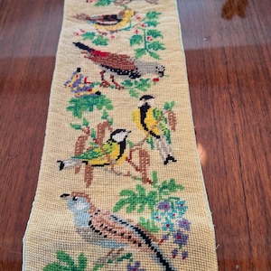 May include: A long, narrow, hand-stitched tapestry featuring colorful birds perched on branches with leaves and berries. The birds are depicted in various colors, including yellow, brown, blue, and red, against a beige background.