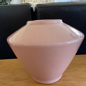May include: A pale pink glass lampshade with a flared, conical shape. The lampshade has a wide top and a narrower base, with a smooth, glossy finish. It is sitting on a wooden surface.