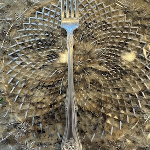 May include: A silver fork with an ornate handle. The fork is resting on a clear glass plate with a decorative pattern.