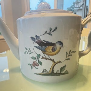 May include: White ceramic teapot with a lid and a curved handle. The teapot features a detailed illustration of a bird perched on a branch with leaves and berries. The bird has brown, blue, and yellow feathers.