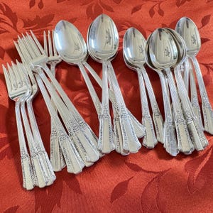 May include: A collection of vintage silverware, including forks and spoons, arranged on a red patterned tablecloth. The silverware has a shiny, reflective surface and ornate detailing on the handles. The spoons have an oval bowl shape.