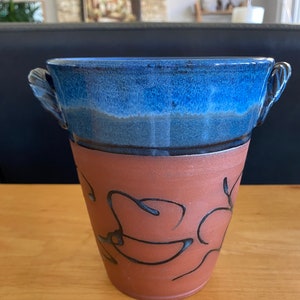 May include: A blue and brown ceramic vase with two handles and a floral design. The vase is glazed in a blue and brown color scheme and has a rustic look.