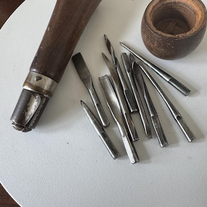 May include: A collection of antique woodworking tools, including a wooden handle with a metal ferrule, various metal chisels, and a small wooden cup. The tools are arranged on a white surface.
