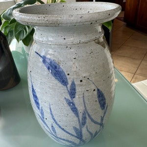 May include: A white ceramic vase with a blue floral design. The vase has a wide mouth and a rounded body. It is decorated with a pattern of blue leaves and stems.