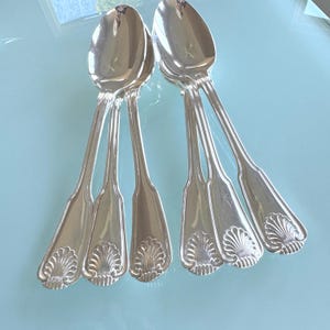 May include: A set of five polished silver-toned spoons featuring a classic fiddle-back handle design with a decorative shell motif at the base of each handle. Each spoon bowl is smooth and reflective, showing a subtle engraved letter K.
