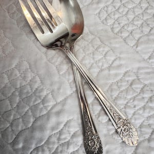 May include: A silver-toned metal fork and spoon crossed over one another. Both pieces of cutlery feature a polished finish and an intricate floral embossed pattern on the handles, with a scalloped edge detail at the base of each handle.