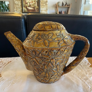 May include: A brown ceramic teapot with a floral design and a woven lid. The teapot has a wide spout and a handle.