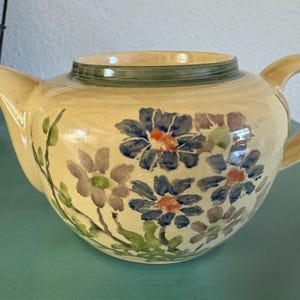 May include: Cream-colored ceramic teapot with a curved spout and handle. The teapot is decorated with hand-painted blue, purple, and orange floral designs. A green band encircles the rim. The teapot sits on a teal surface.