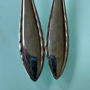 May include: Two silver spoons with rounded ends and decorative details. The spoons are placed side by side, with the rounded ends facing each other.