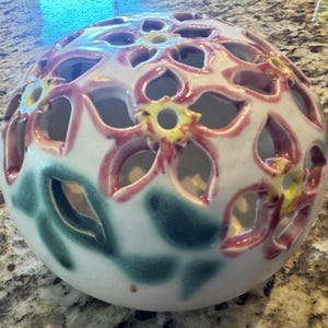 May include: A ceramic sphere with a floral design. The sphere is white with cut-out flower shapes outlined in red and yellow. Green leaf shapes are painted on the bottom of the sphere.
