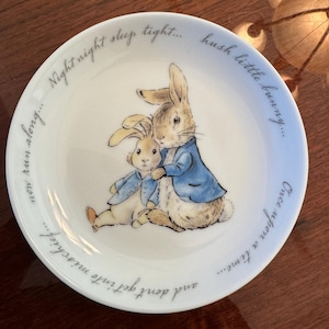 May include: A white ceramic mug with a black handle and the text "But First, Coffee" in a bold, black font. The mug is set against a blurred background, suggesting a cozy setting.