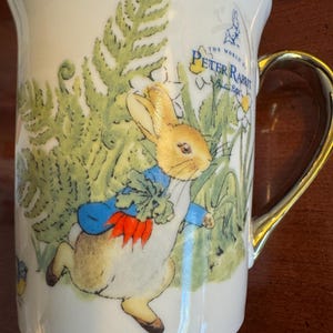 May include: White ceramic mug featuring a colorful illustration of Peter Rabbit from Beatrix Potter's story. The mug has a gold-colored handle and the words "The World of Peter Rabbit" printed on the side. The design includes flowers and foliage.
