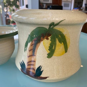 May include: A ceramic planter with a beige background and a painted green palm tree with a yellow sun. The planter has a slightly flared rim.