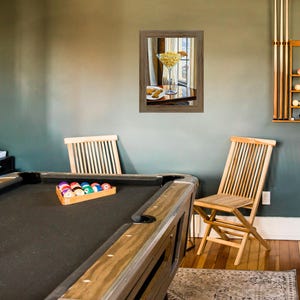 May include: A green felt pool table with a wooden frame and a set of billiard balls in a wooden rack. Two wooden chairs are in the background, one with a framed picture of a martini glass with olives hanging above it.