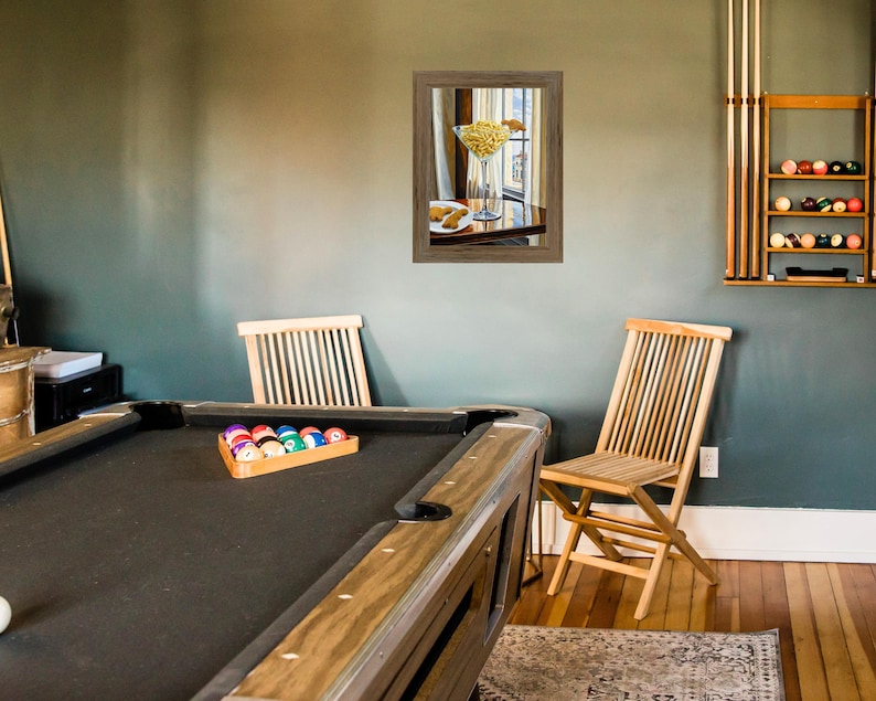 May include: A green felt pool table with a wooden frame and a set of billiard balls in a wooden rack. Two wooden chairs are in the background, one with a framed picture of a martini glass with olives hanging above it.