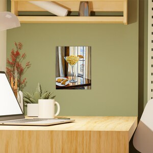May include: A painting of a glass martini glass filled with yellow french fries, sitting on a wooden table with a plate of cookies in the background. The glass is on a white tablecloth and the table is in front of a window.