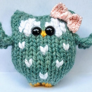 May include: Hand-knitted owl figurine in shades of teal, white, and yellow. The owl has large black eyes, a yellow beak, and a pink bow. The body is decorated with white heart-shaped accents. A charming, handmade decorative item.